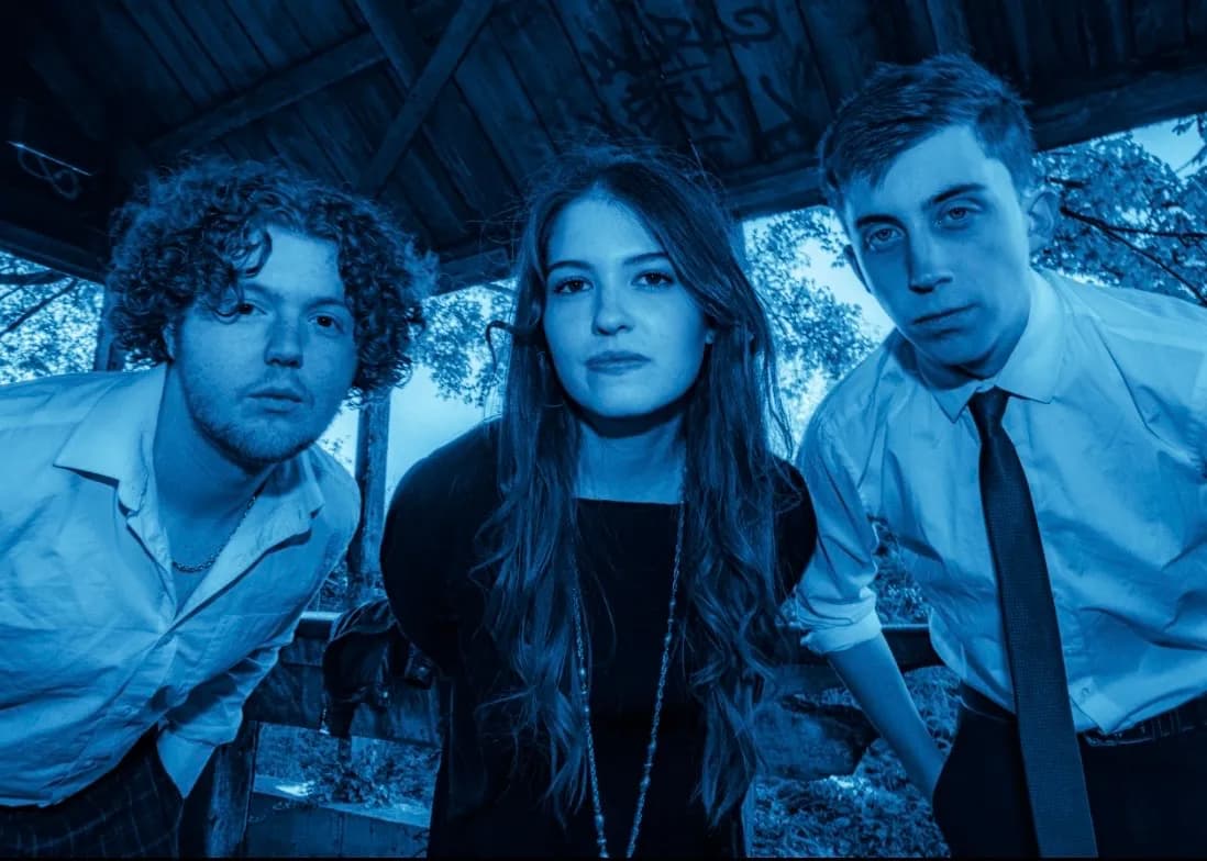 Three young adults, two men and one woman, with serious expressions, are illuminated by blue light in an outdoor setting with trees and a wooden structure in the background.