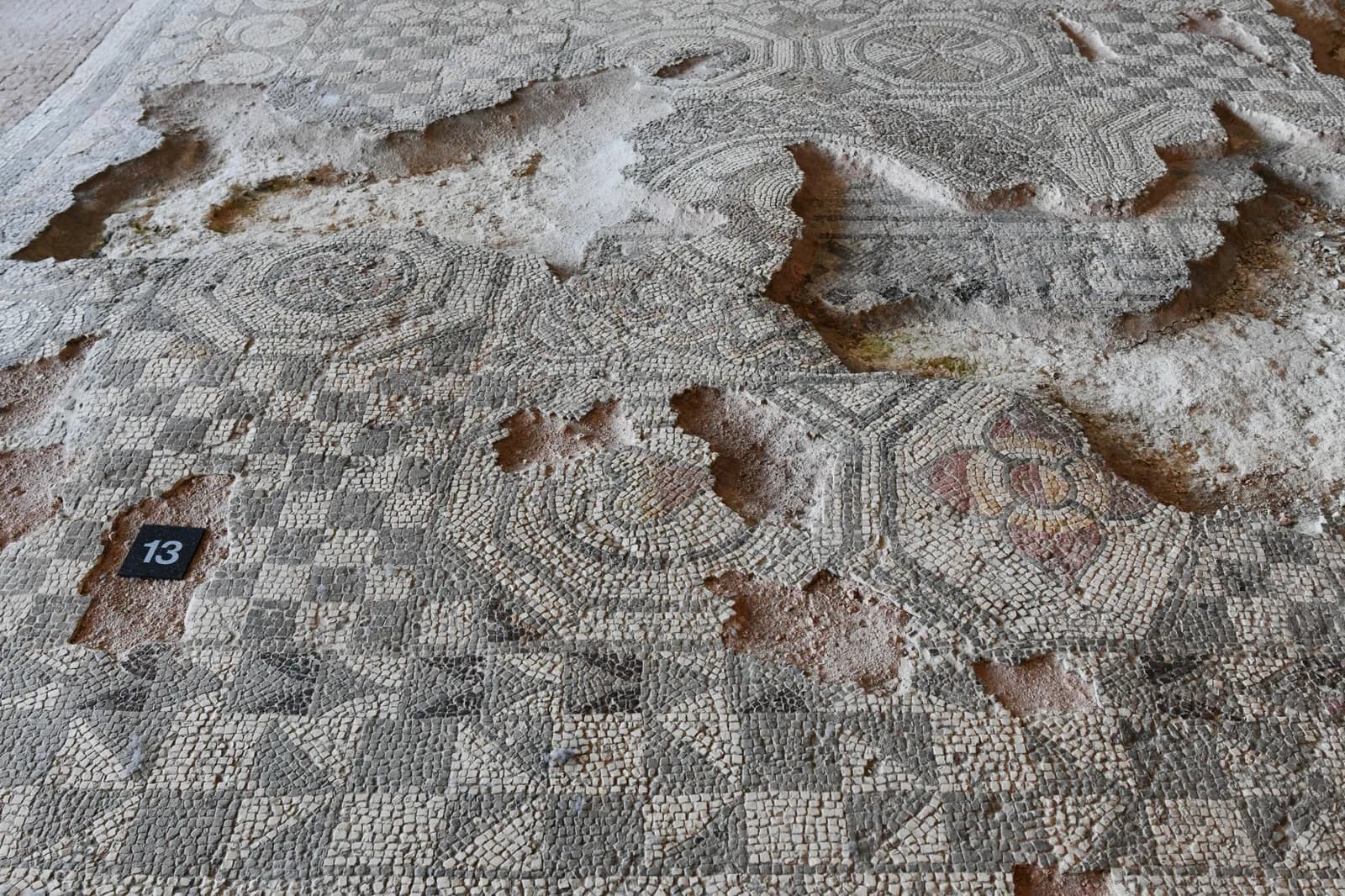 A close-up view of a damaged ancient mosaic floor featuring geometric patterns in varying shades of grey, white, and a hint of red, with sections missing revealing the earthen layer beneath.