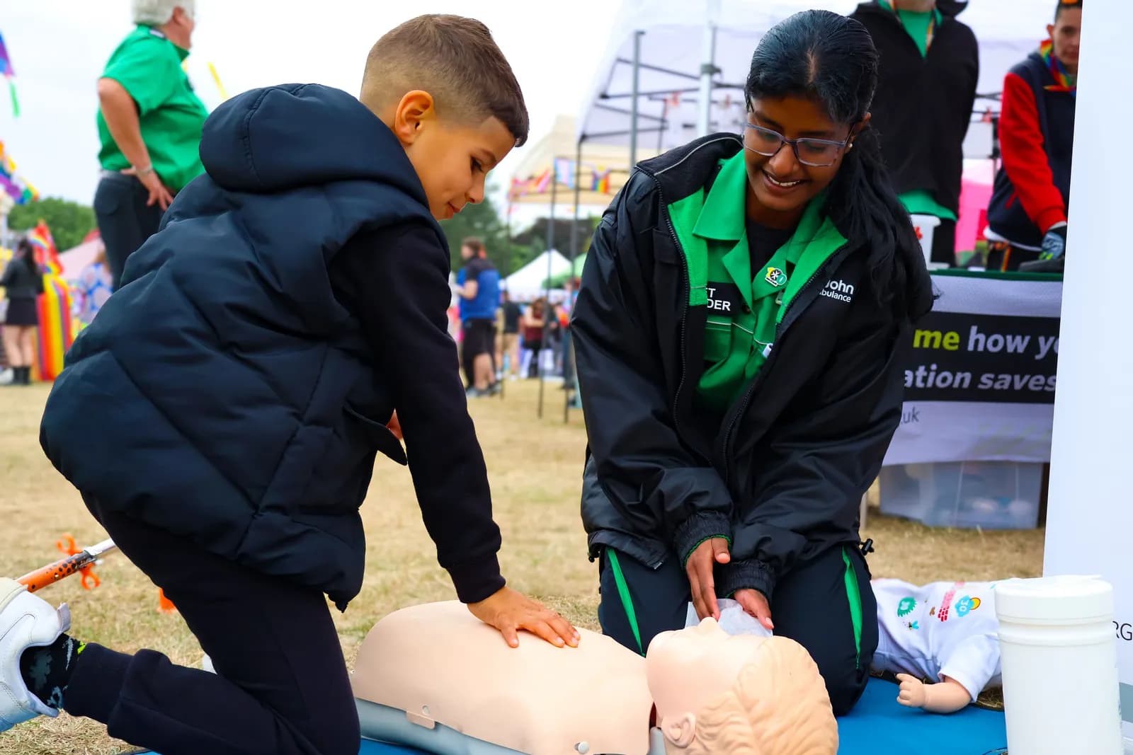 A boy in a dark vest practices CPR on a manikin with a woman in a St. John Ambulance uniform demonstrating beside him.