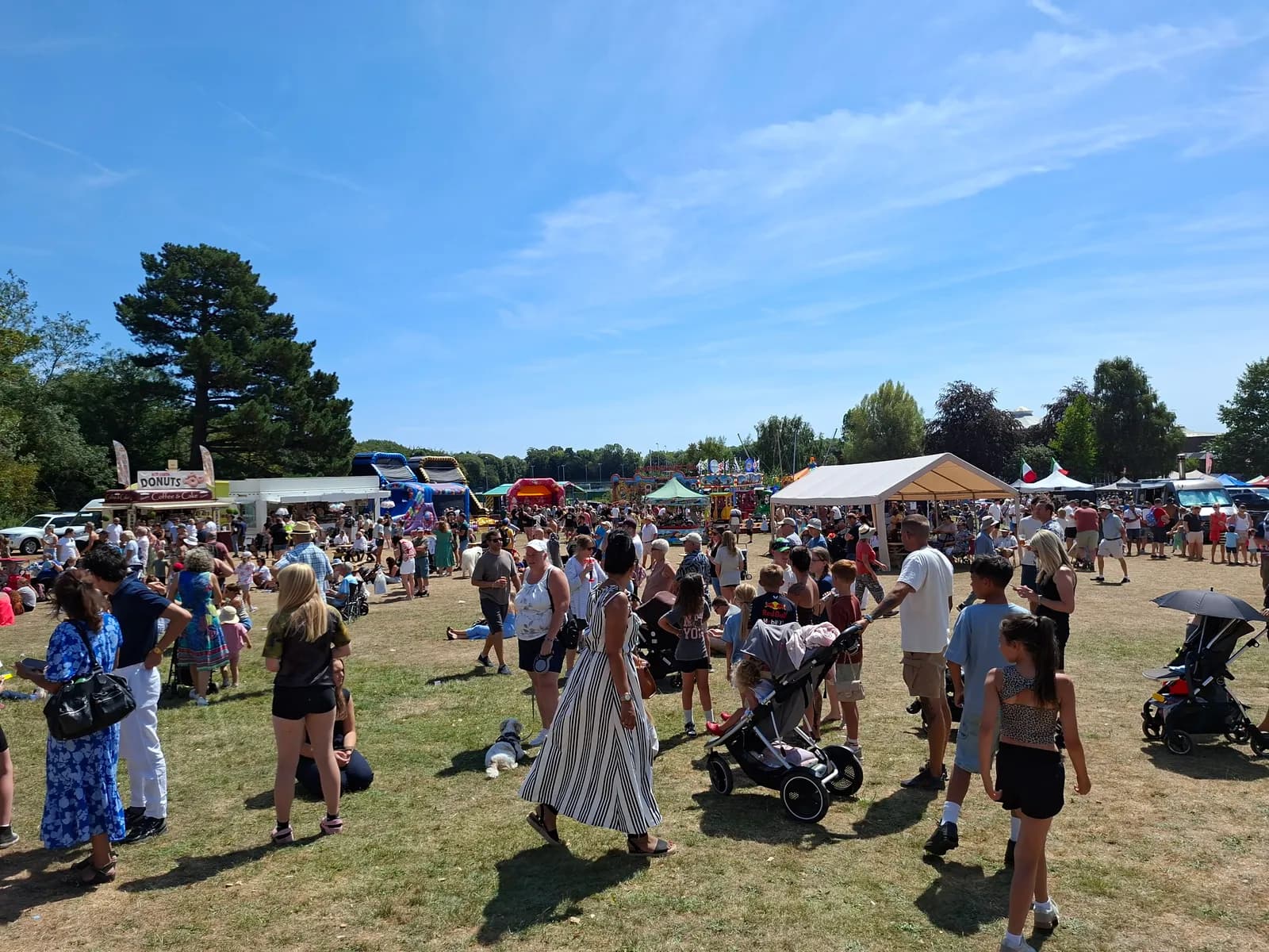 A wide outdoor shot captures a bustling fairground under a bright blue sky, with numerous people walking around and various stalls visible in the background, including a "Donuts" stand.