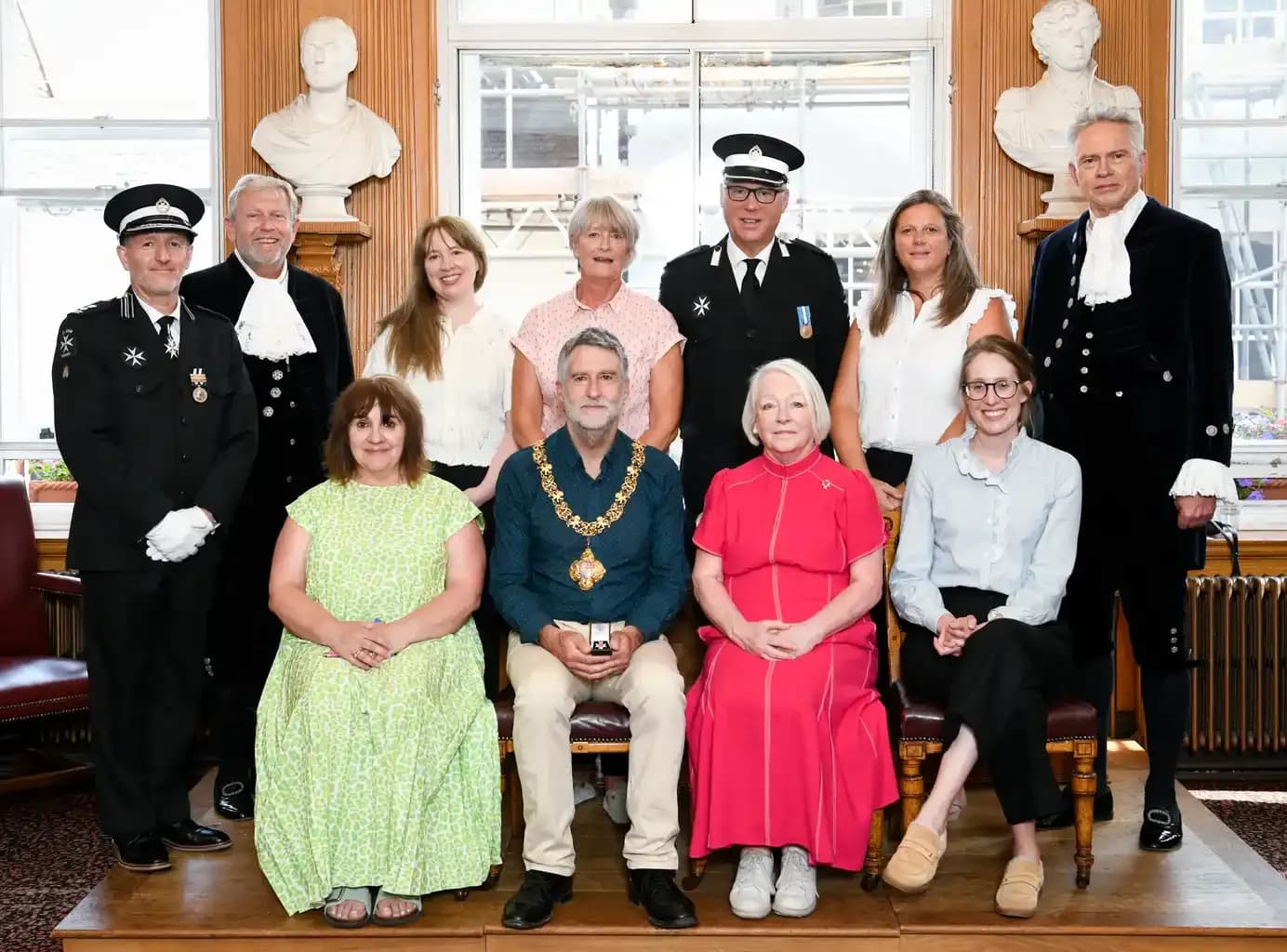 A large group of people posing for a formal photo, with some seated in the front row and others standing behind them. Several individuals are wearing ceremonial attire, including a man in a black uniform with medals and a hat, and a man in a dark blue robe with white ruffles. Two individuals in the