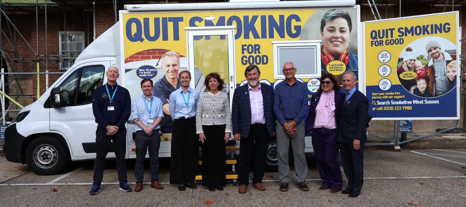 A group of ten people, some smiling at the camera, stand in front of a white truck with a yellow sign that reads "Quit Smoking for Good" and features images of people.